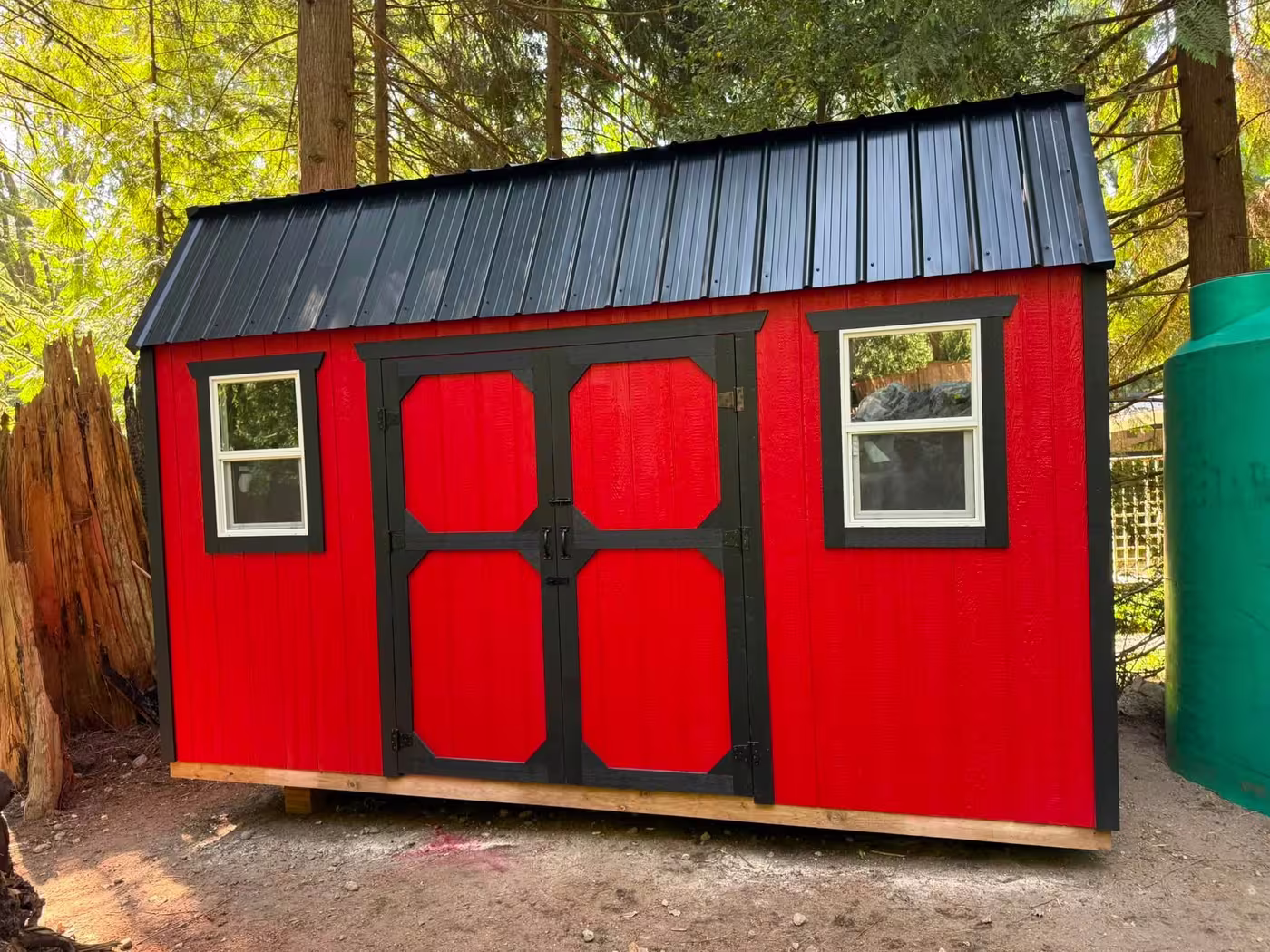 Red barn-style shed with a black roof, black double doors and two windows among trees