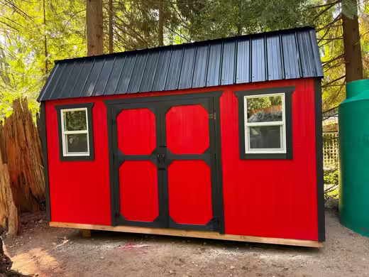 Red barn-style shed with a black roof, black double doors and two windows among trees